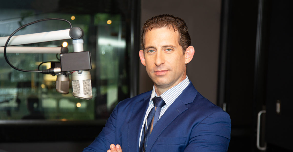 Besuited disability lawyer Sivan Tumarkin stands next to a microphone, with a confident smile and crossed arms