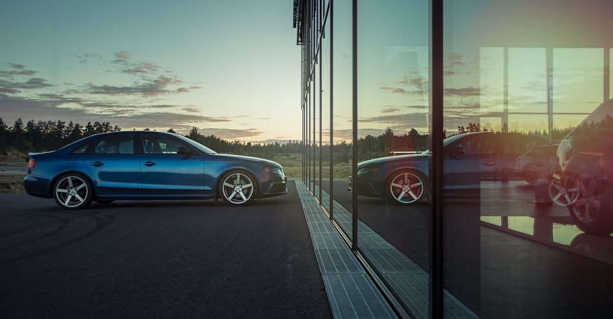 A blue car is parked in front of a reflective glass facade, with a setting sun in the background. Ford Canada employees are entitled to full severance when they lose their job.