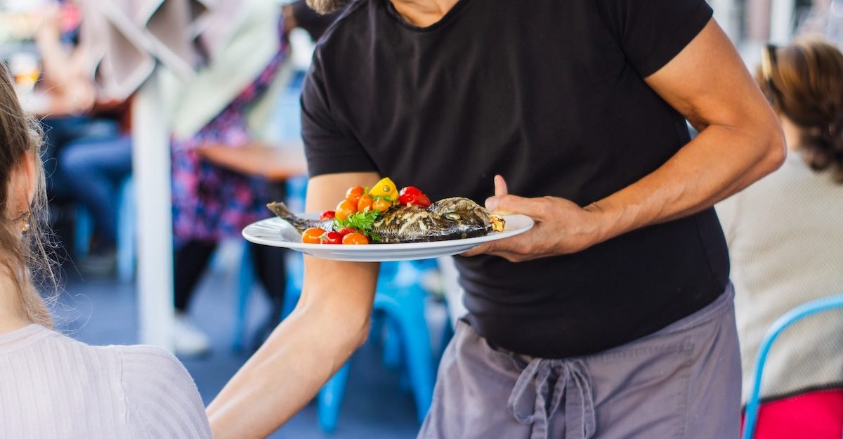 A restaurant server in a black t-shirt delivers a plate of food to a customer's table.