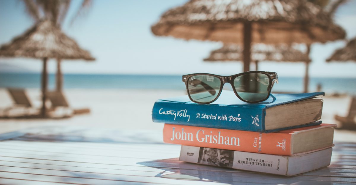 Three books with a pair of sunglasses on top sit on a table with beach and umbrellas in the background.