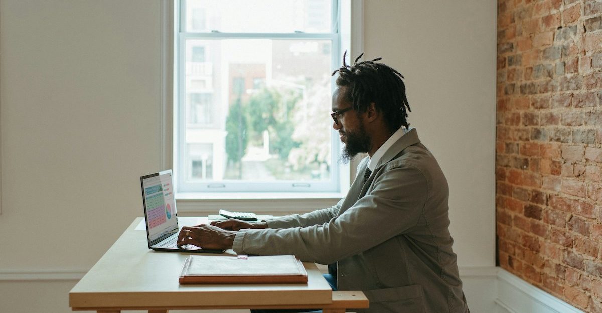 A man sitting at a wooden table in a room in his home, working remotely on his laptop.