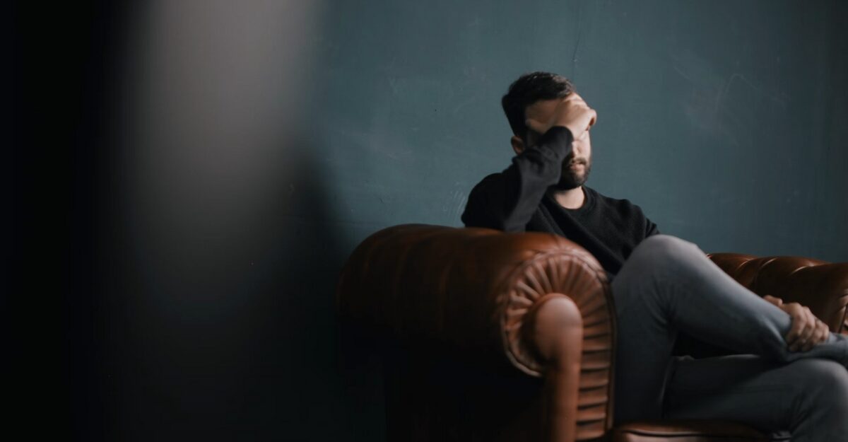 A person sitting on a brown couch with their hand over their face. (Photo: Nik Shuliahin / Unsplash)