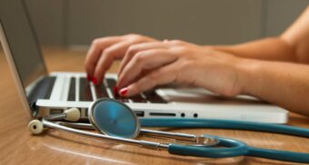 A person uses a laptop with a stethoscope beside them. (Photo: National Cancer Institute / Unsplash)