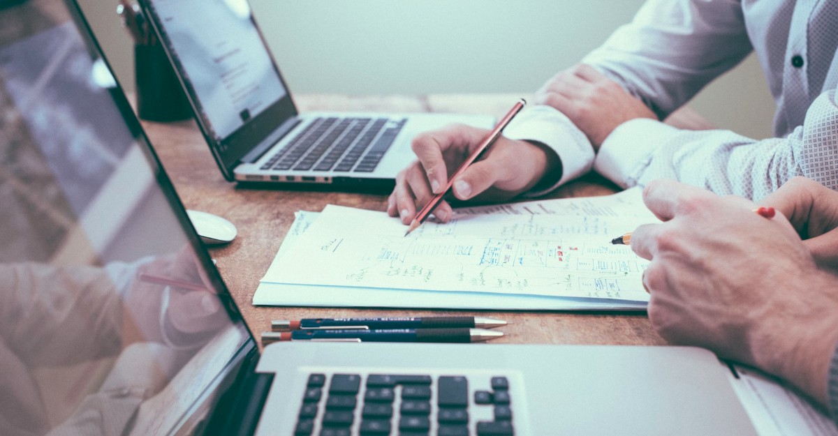 Two people looking at a piece of paper between laptops. (Photo: Scott Graham / Unsplash)